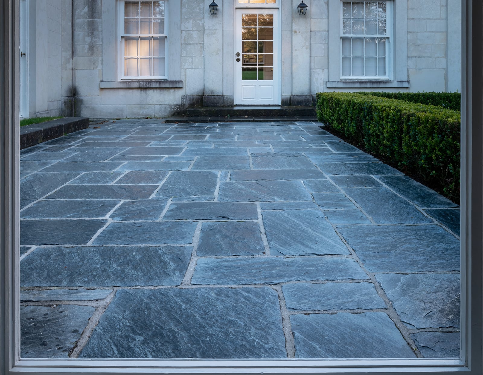 The view from inside a Georgian house looking out onto a Kilkenny Blue Limestone patio with large random-pattern flags and lime mortar joints, clipped box hedge to one side, soft evening light.